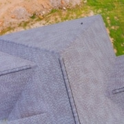 Precision in Action123 aerial shot of a finished gray shingle roof with clean ridge lines, next to construction dirt and lawn.