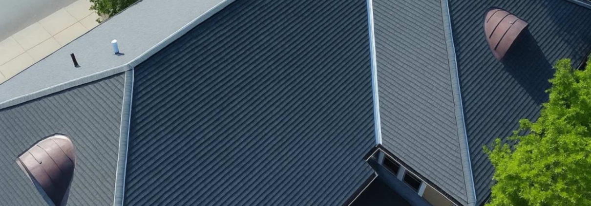 Aerial view of a large roof installation featuring dark gray standing seam metal panels and two distinctive semicircular copper-toned dormer vents.