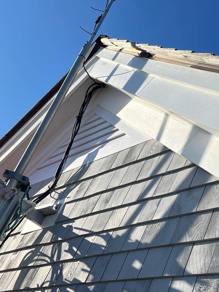 2023-12-16 14 33 22Z-lq85rm3foxuw6ro9 a house gable with weathered gray shingle siding, white trim, a triangular vent, and electrical wires running along a metal pole in the sunlight.