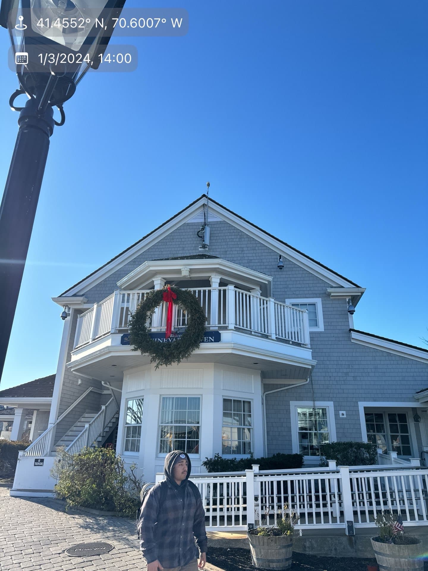 2024-01-03 19 00 49Z-lqy58vqkvrykhkyu Gray shingle building with a balcony, a large wreath, and a person walking in the foreground.