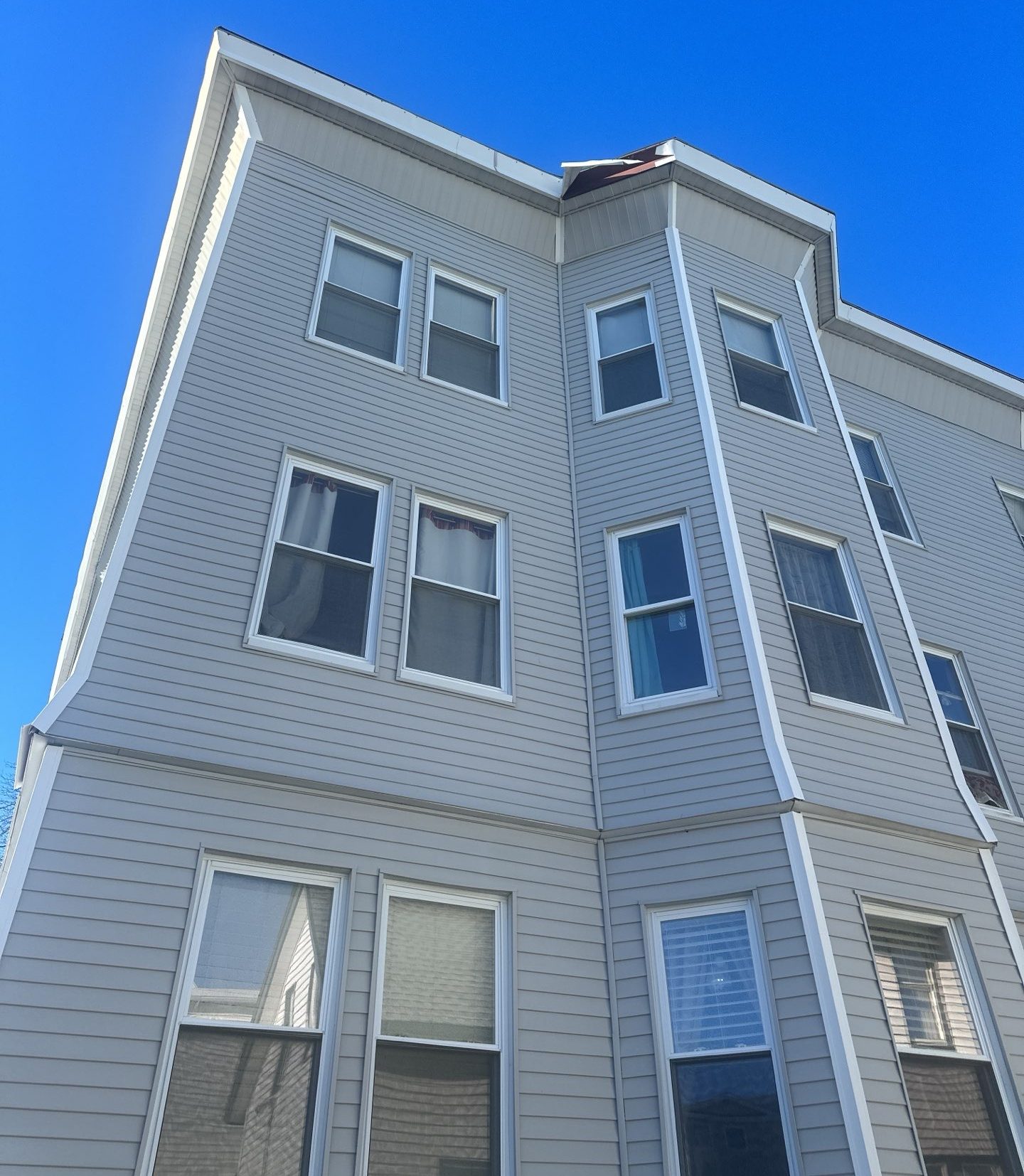 2024-01-05 17 56 31Z-lr0xtwsohdc9am92 Three-story building with light gray siding and many white-trimmed windows against a clear blue sky.