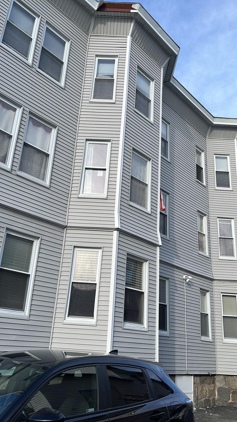 2024-01-18 15 58 11Z-lrjebsw1u9k1fkeo Corner of a multi-story building with gray vinyl siding and white trim over a stone foundation.
