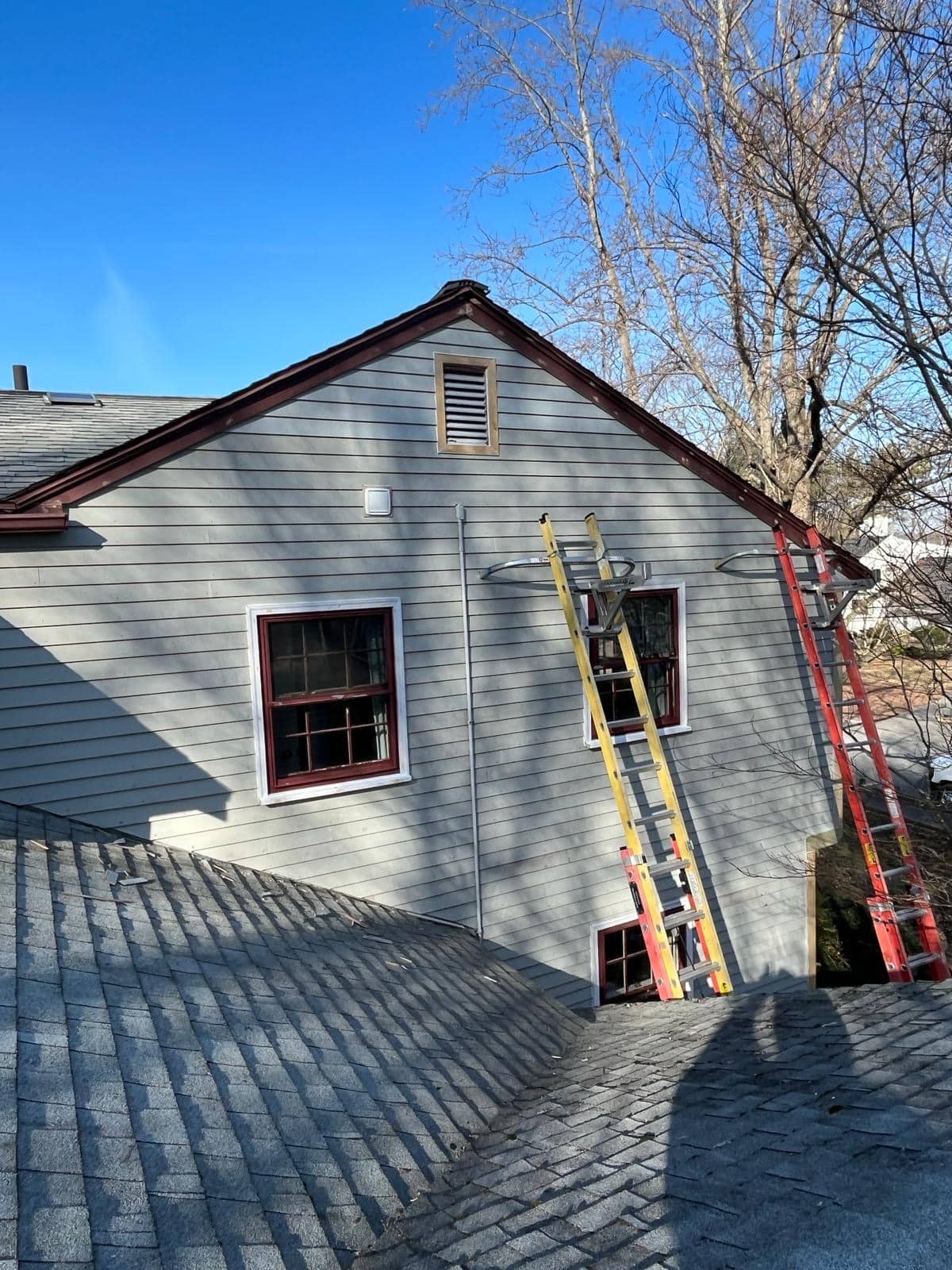 2024-02-26 21 59 50Z-lt3hf3focg0n765j Gable end of a light gray house with two ladders and red-trimmed windows, viewed from a shingled roof.