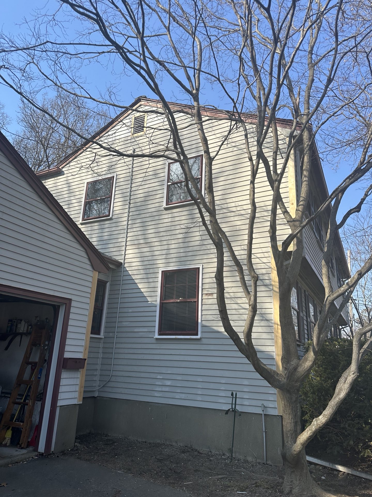 2024-02-27 21 07 17Z-lt4uzdsyd1v5e10i Side of a light gray house undergoing trim replacement next to a garage, with a bare tree in the foreground.