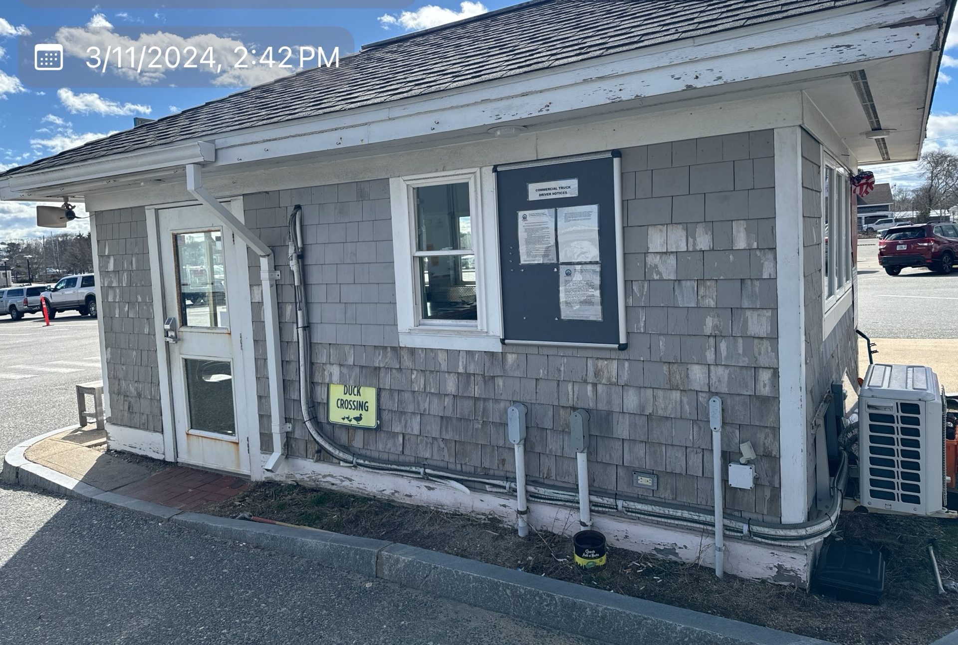 2024-03-11 18 42 51Z-ltnajphc5oszcaex Small gray shingle building with a window, posted notices, and a "Duck Crossing" sign.