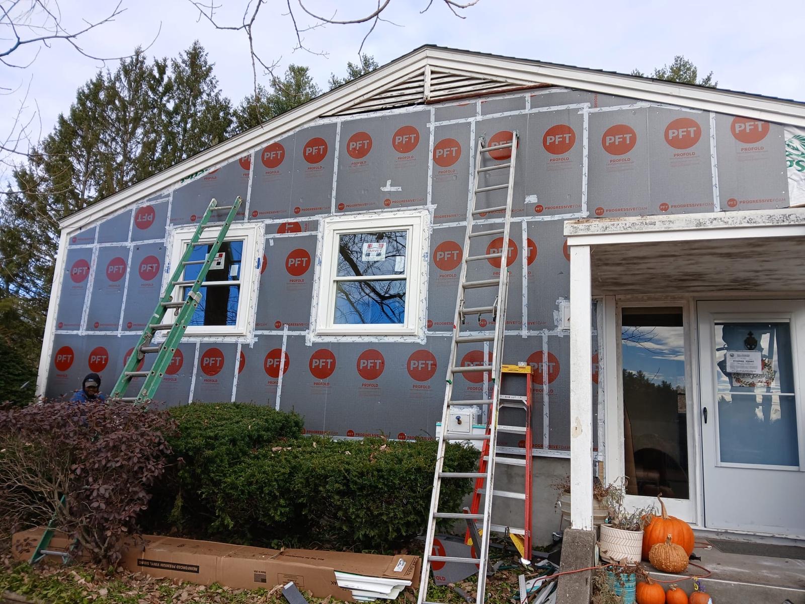 2024-11-20 23 53 42Z-m3qjhu0ttwbjlmdg House wall covered in dark gray insulation board with orange PFT logos, two new windows, and ladders in place for siding installation.