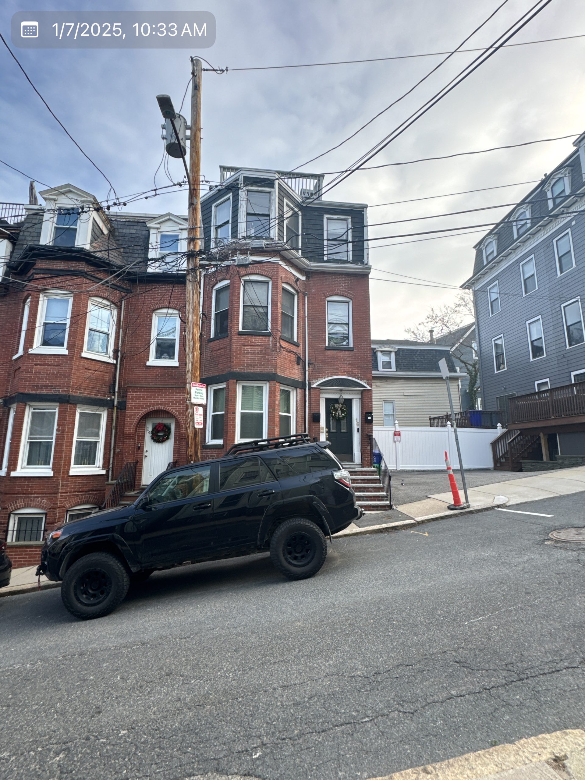 2025-01-07 15 33 54Z-m5mmrzh46g159qef Street view of a three-story brick apartment building with bay windows and a mansard roof on an overcast day.