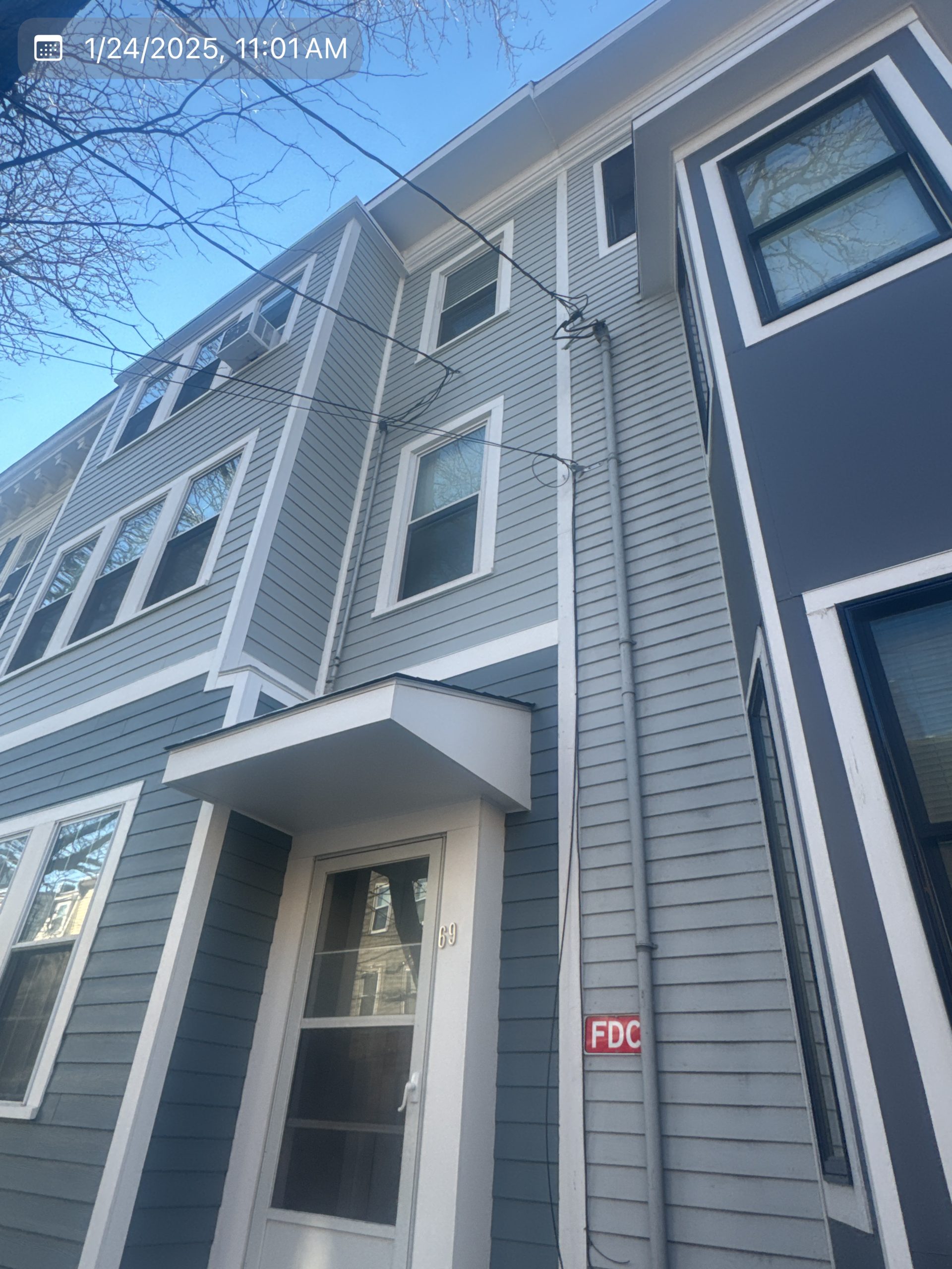 2025-01-24 16 01 28Z-m6ay8wdi2ove36p0 Looking up at the tall, narrow side of a house with different shades of gray siding against a bright blue sky.