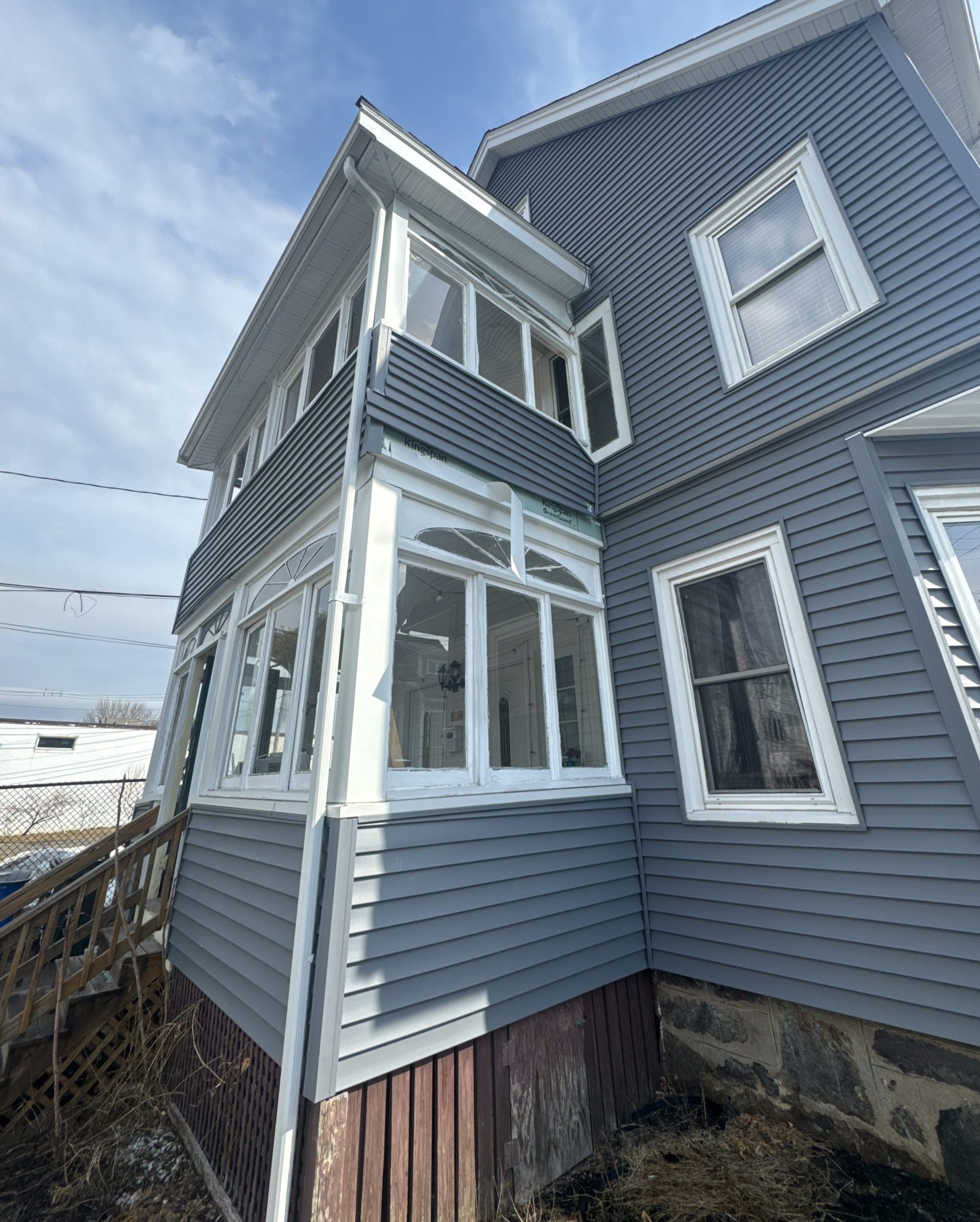 2025-02-27 16 19 40Z-m7njva89zj4cjt5z Low-angle exterior view of a house corner with new dark gray siding contrasting with an old, enclosed porch section with many windows and white trim.