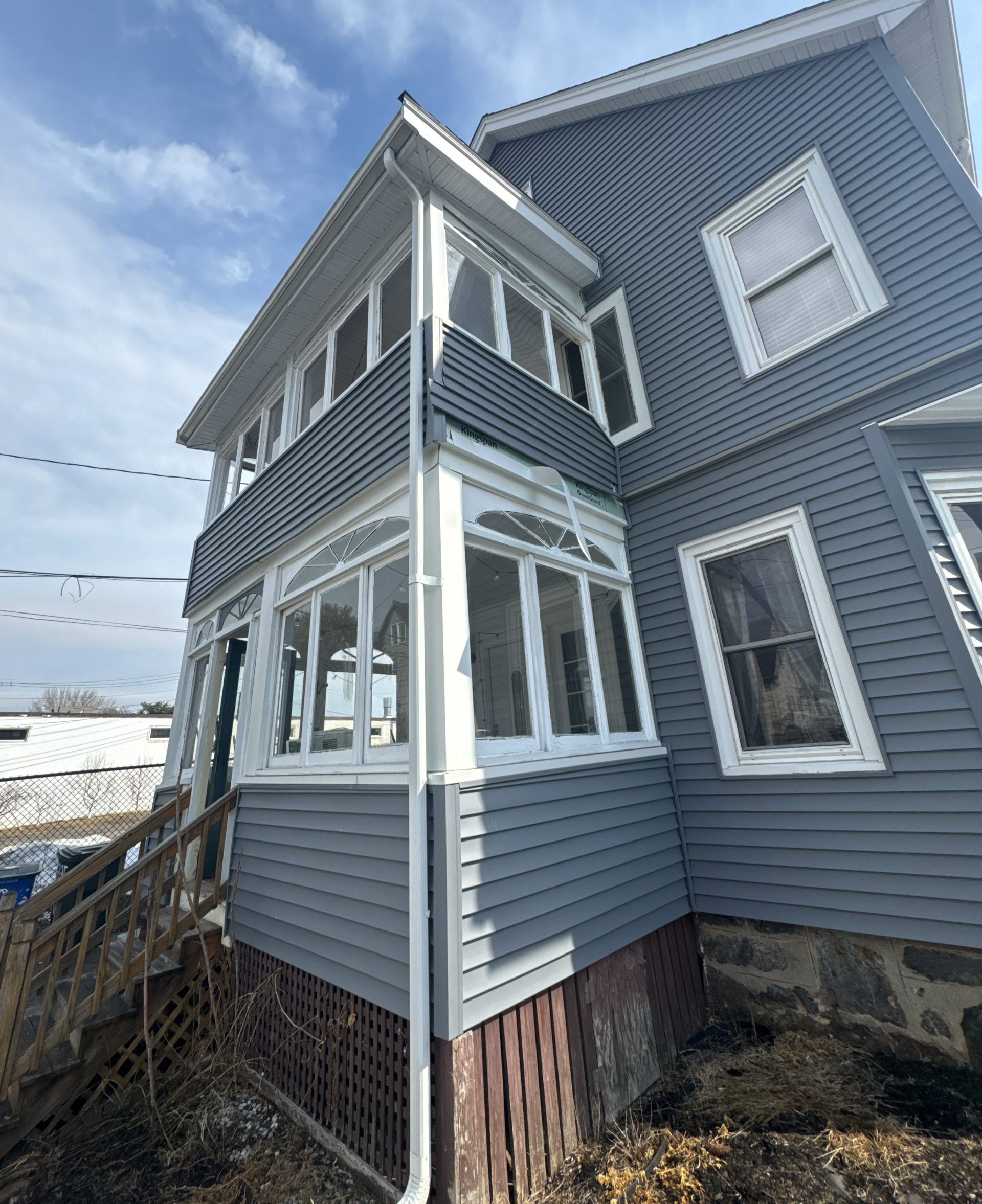 2025-02-27 16 20 20Z-m7njw4t8m6b1s8v6 Corner of a house with new gray siding, an enclosed multi-window porch with white trim, and old wooden stairs leading up to the entrance.
