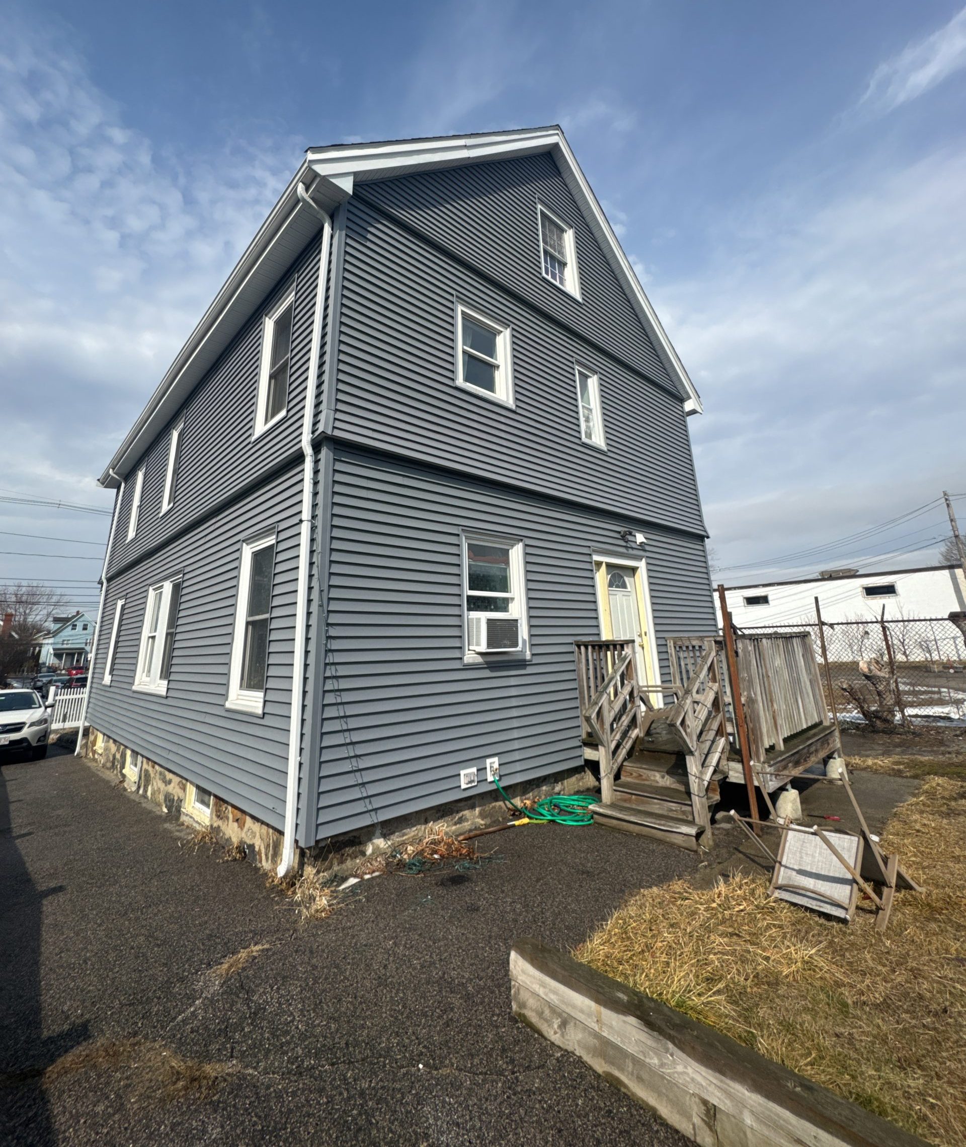 2025-02-27 16 21 06Z-m7njx4otm42xprhv Two-story house with new gray vinyl siding, white trim, and a worn wooden deck and stairs leading to a back entrance under a partly cloudy sky.