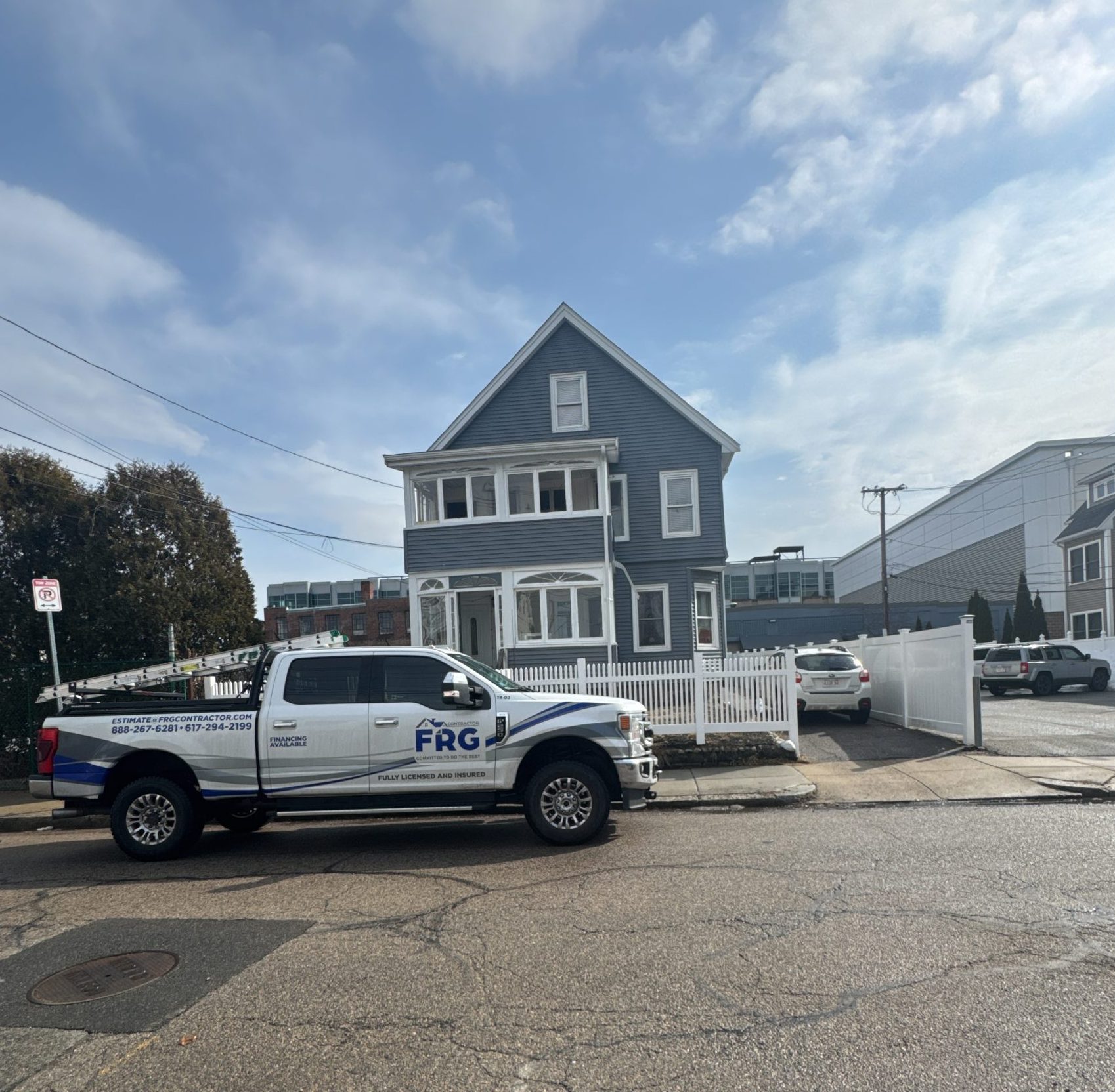 2025-02-27 16 22 26Z-m7njytrw8u17a71i A white contractor's pickup truck with an FRG logo and ladders parked in front of a blue two-story house with new siding and a white fence.