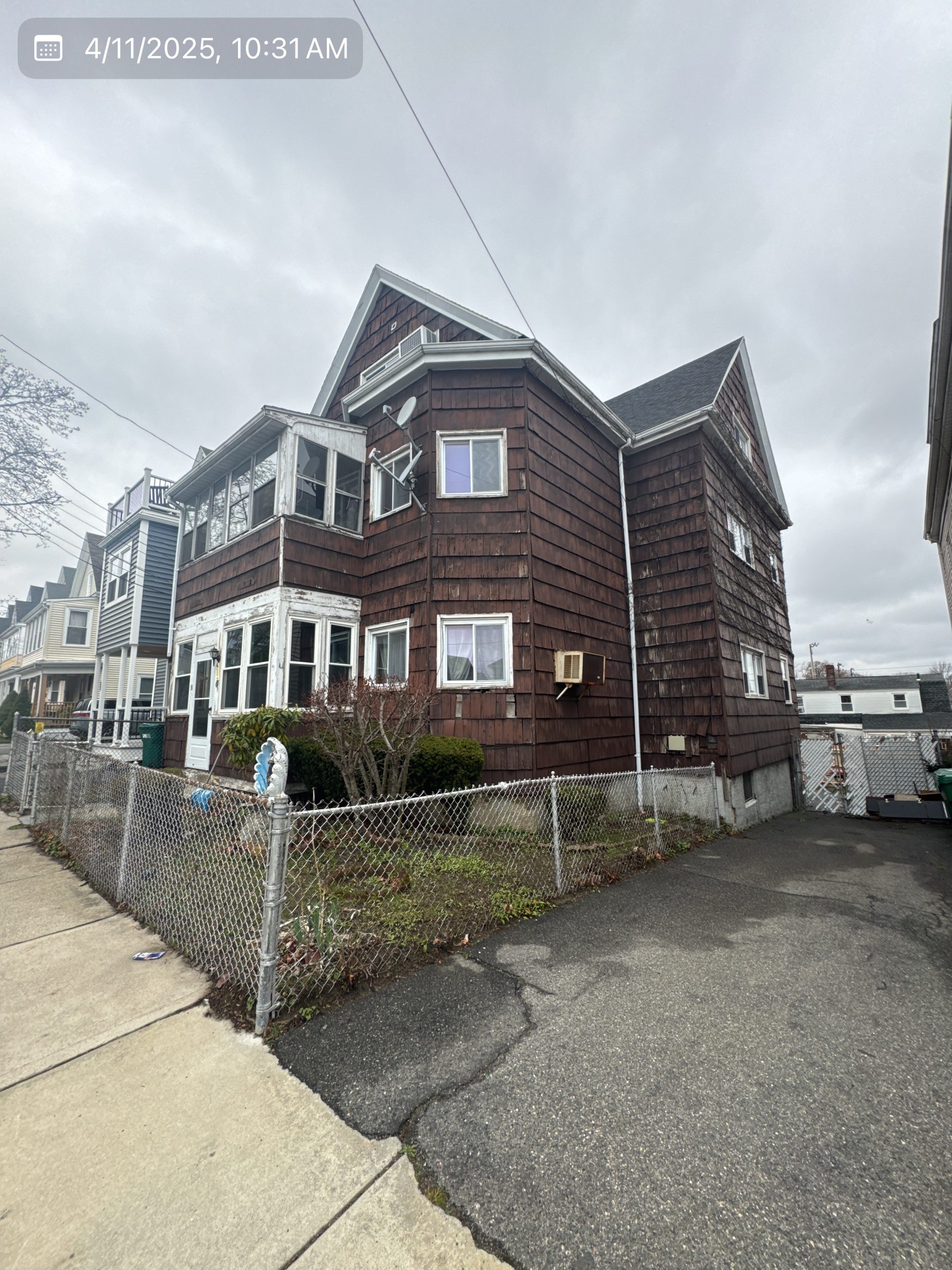2025-04-11 14 31 38Z-m9cvxytqfm9r8ab4 Two-story house with dark brown shingle siding, a worn-out enclosed porch, and white-framed windows, viewed from the side street under a cloudy sky.