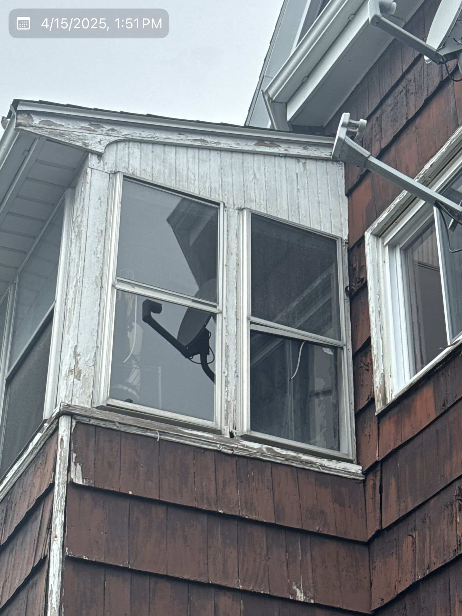 2025-04-15 17 51 14Z-m9isu2ezvumuwker Exterior of a house showing weathered brown shingle siding and a bay window area with peeling white paint and old windows.