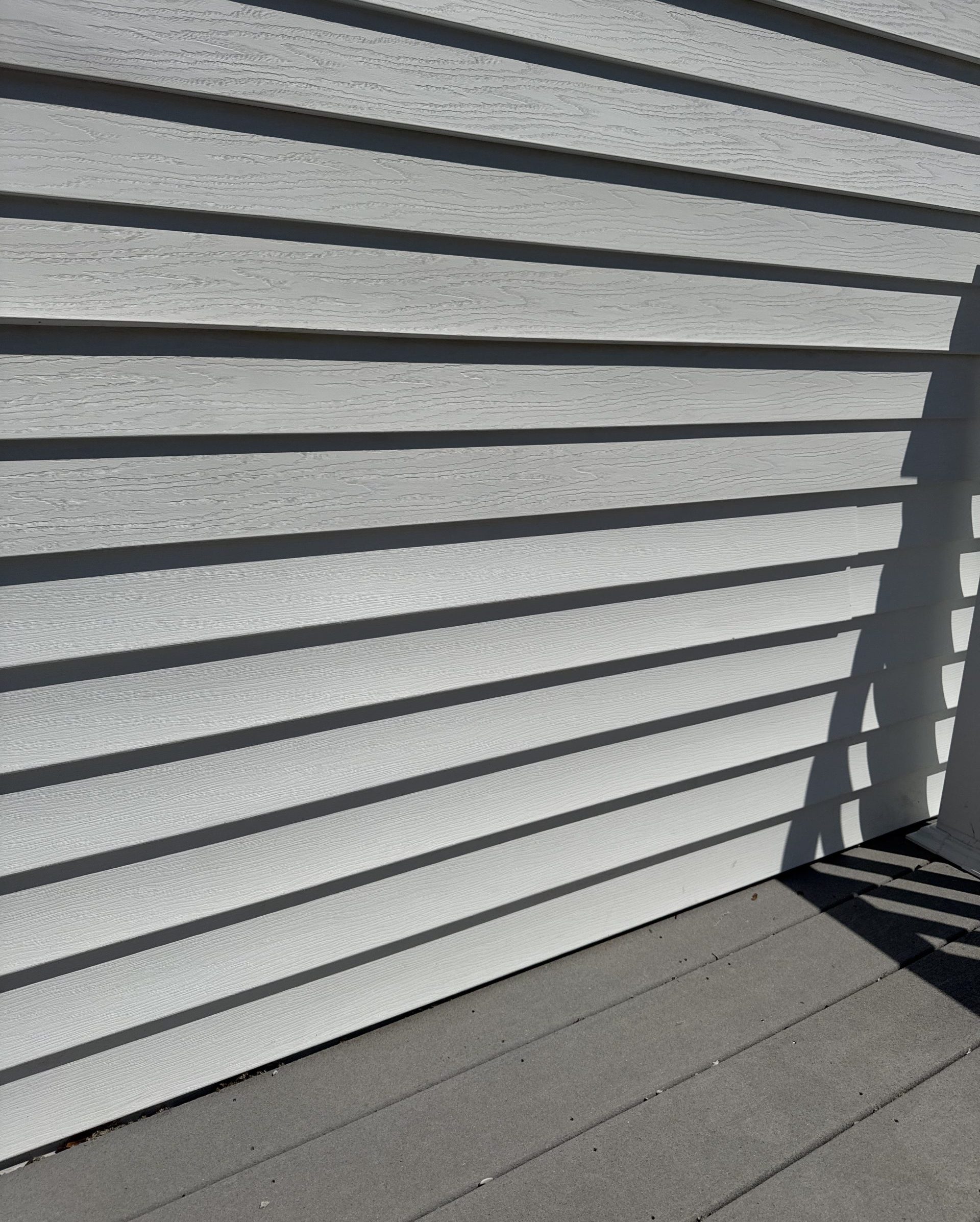 2025-04-21 17 16 18Z-m9rc88y19q82x50d Close-up of white horizontal vinyl siding with deep shadows from direct sunlight, meeting a gray composite deck floor.