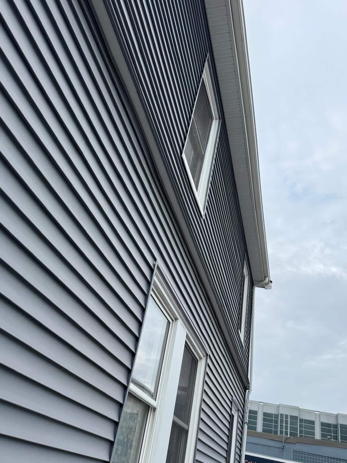 2025-04-22 12 47 29Z-m9si2f190v23fpiz Angled view of dark gray horizontal vinyl siding and white window frames, stretching toward a cloudy sky.