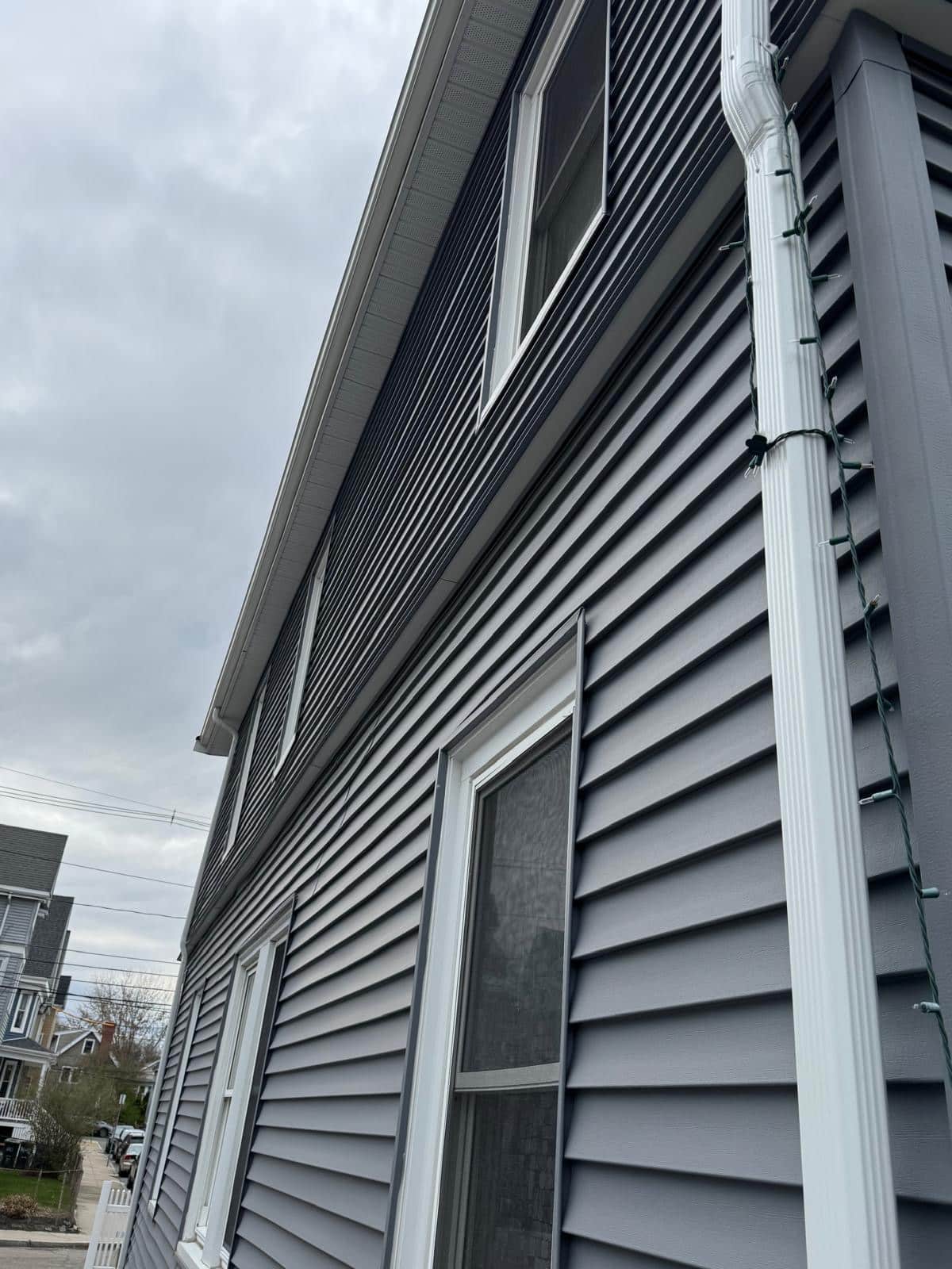 2025-04-22 12 47 29Z-m9si2f1amufez7v10 Side view of a house with dark gray horizontal siding, white gutters and trim, and thin holiday lights strung along the gutter under an overcast sky.