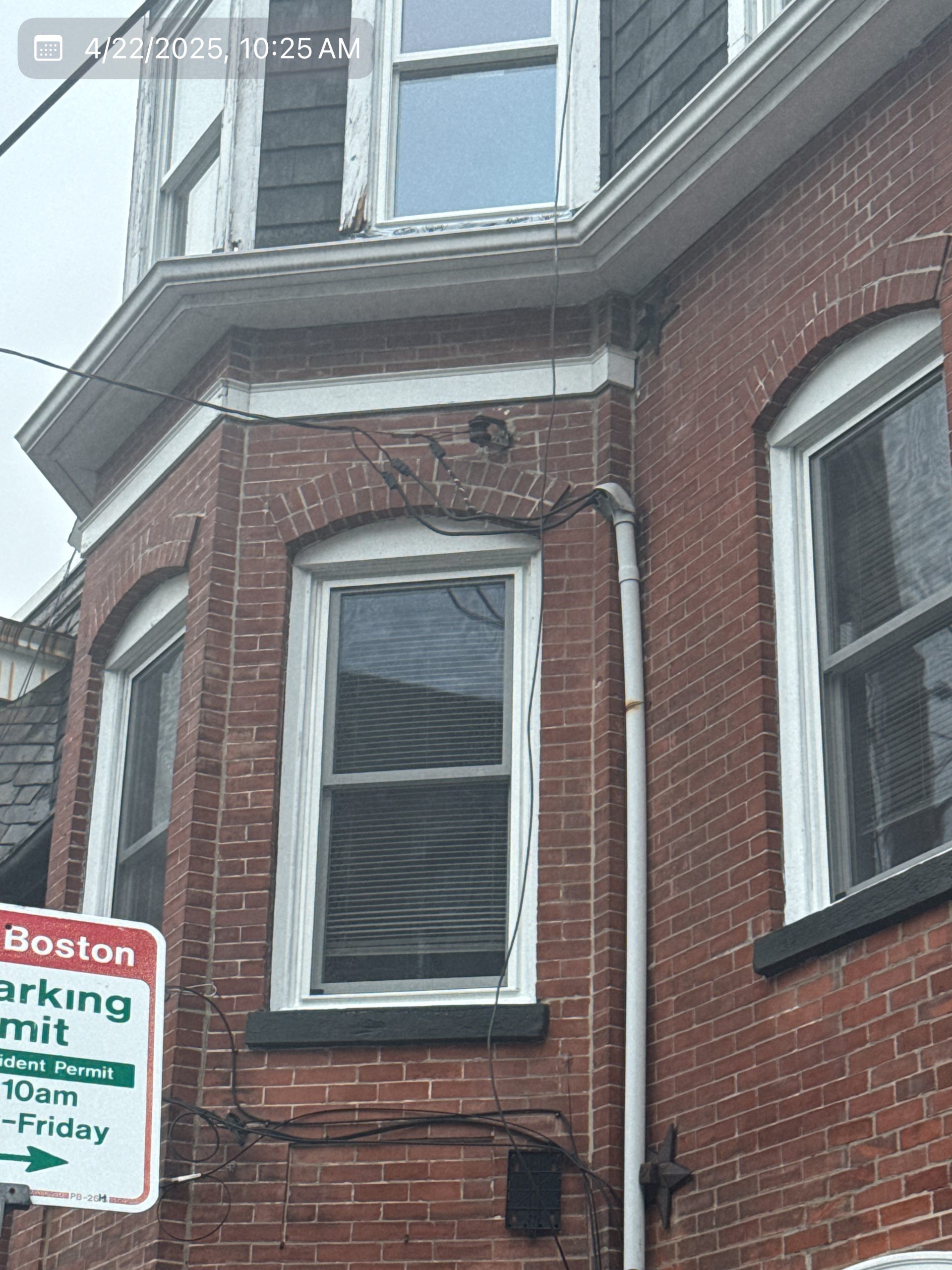 2025-04-22 14 25 49Z-m9slkuyb8vzbsft4 Street view of a three-story brick apartment building with bay windows and a mansard roof on an overcast day.