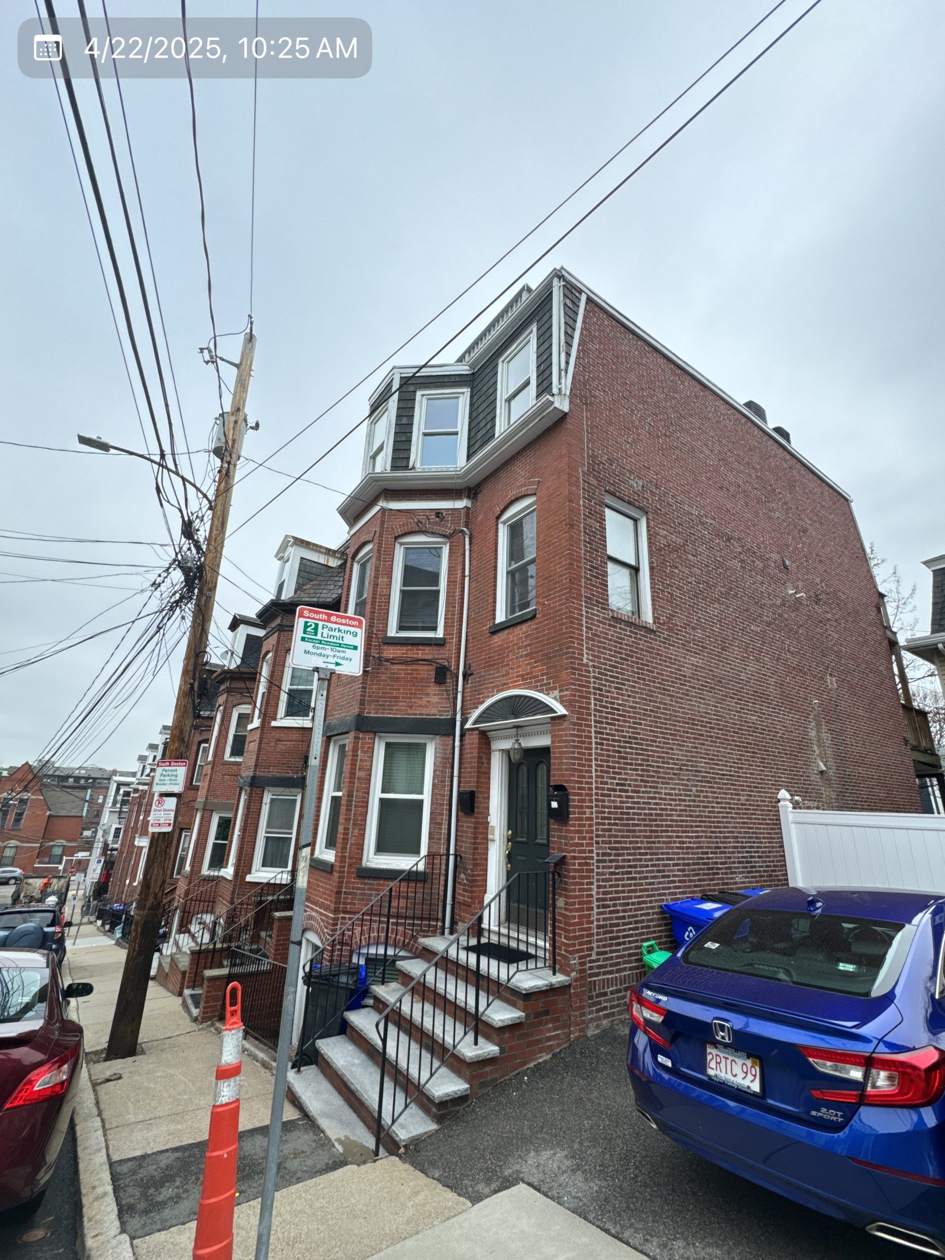 2025-04-22 14 25 57Z-m9sll1aduqkz8bf0 Street view of a three-story brick apartment building with bay windows and a mansard roof on an overcast day.