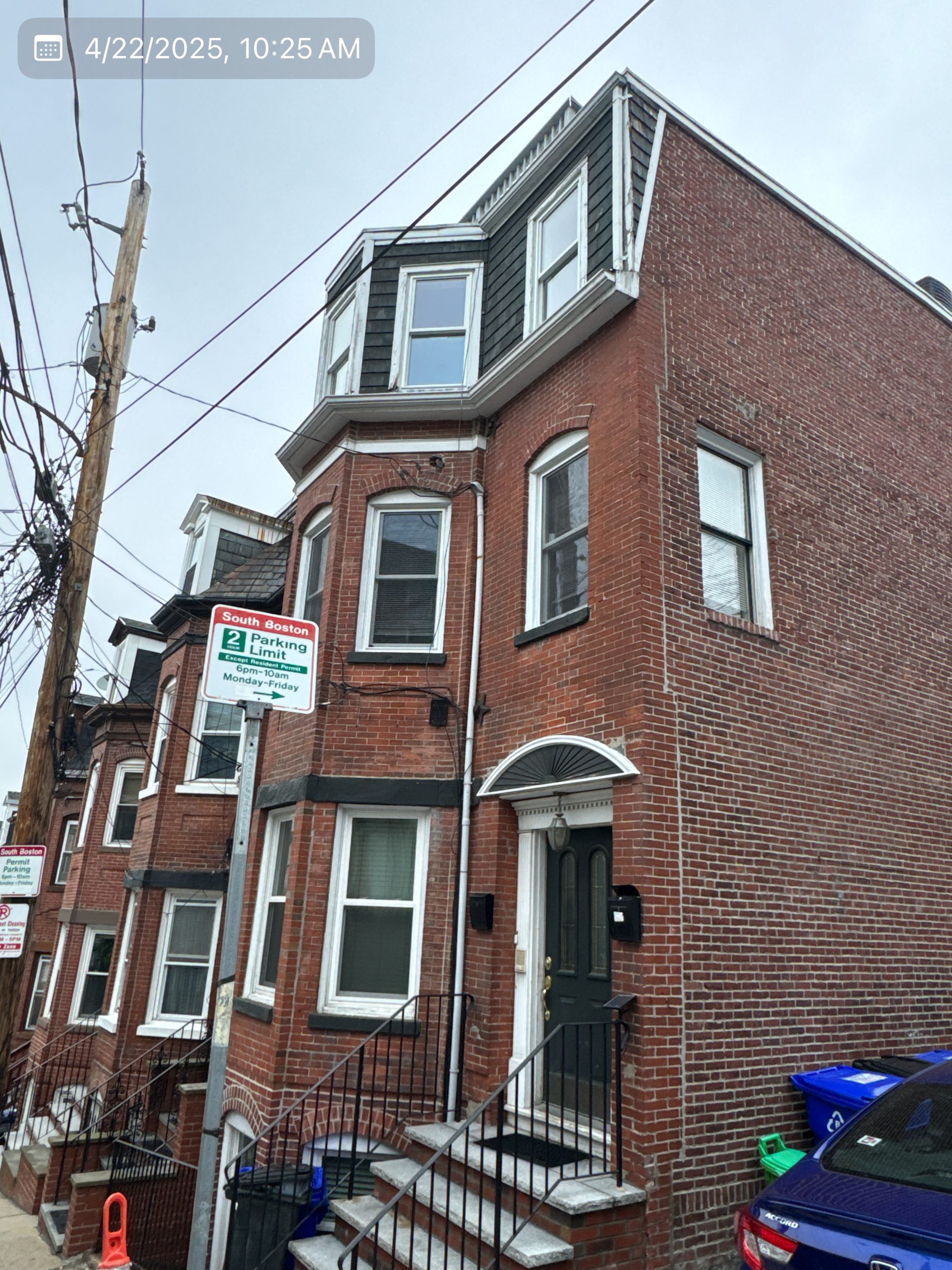 2025-04-22 14 25 59Z-m9sll2hrxkxs3gv8 Street view of a three-story brick apartment building with bay windows and a mansard roof on an overcast day.