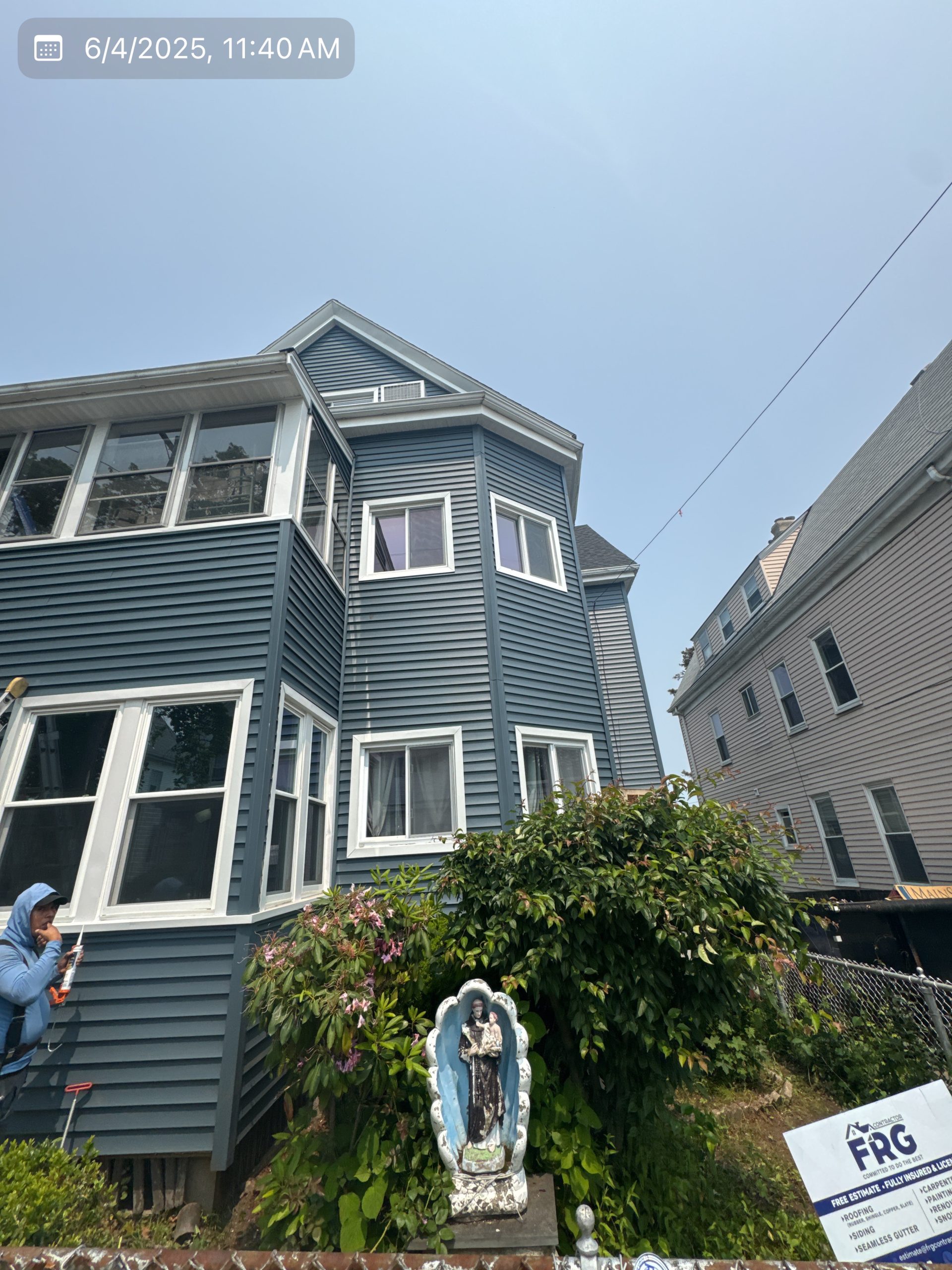 2025-06-04 15 40 05Z-mbi4604txu720hsv Two-story house with new dark blue siding and white trim, featuring a religious statue in the foreground and a worker on a ladder near a window.