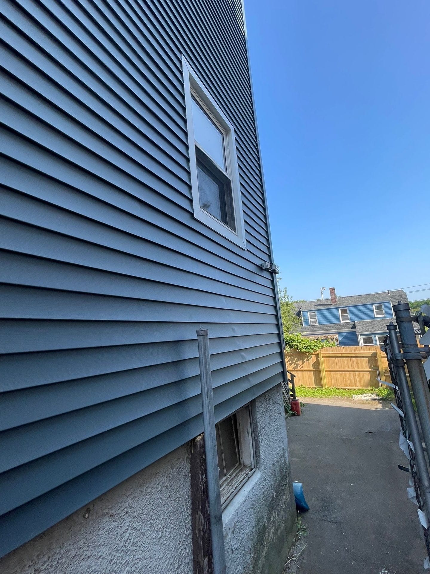 2025-06-05 13 32 32Z-mbjf1tc7enx7rw78 the dark blue horizontal vinyl siding on a house exterior with one vertical window, looking toward a neighbor's yard under a blue sky.