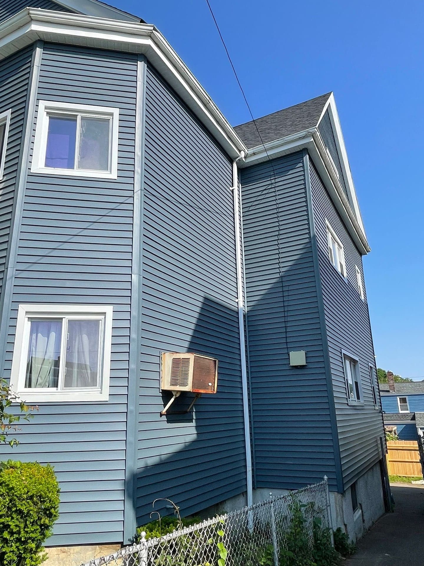 2025-06-05 13 32 32Z-mbjf1tccicgqw7gd Exterior corner of a multi-story building with dark blue horizontal siding, white trim, and a small, rusty air conditioning unit mounted on the wall.