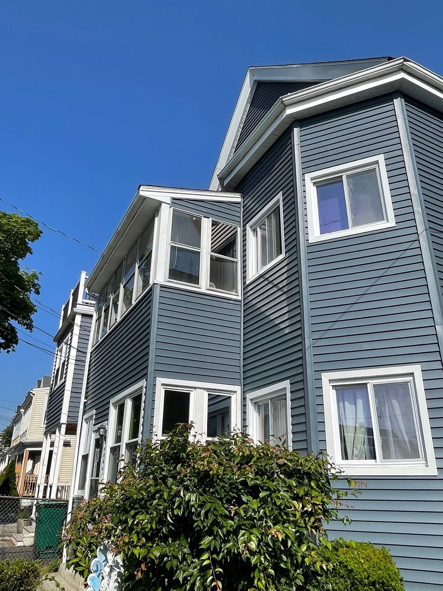 2025-06-05 13 32 32Z-mbjf1tcfex03r4xg Exterior of a multi-story house with dark blue siding, white trim, a bay window section, and a dense green shrub in the foreground.