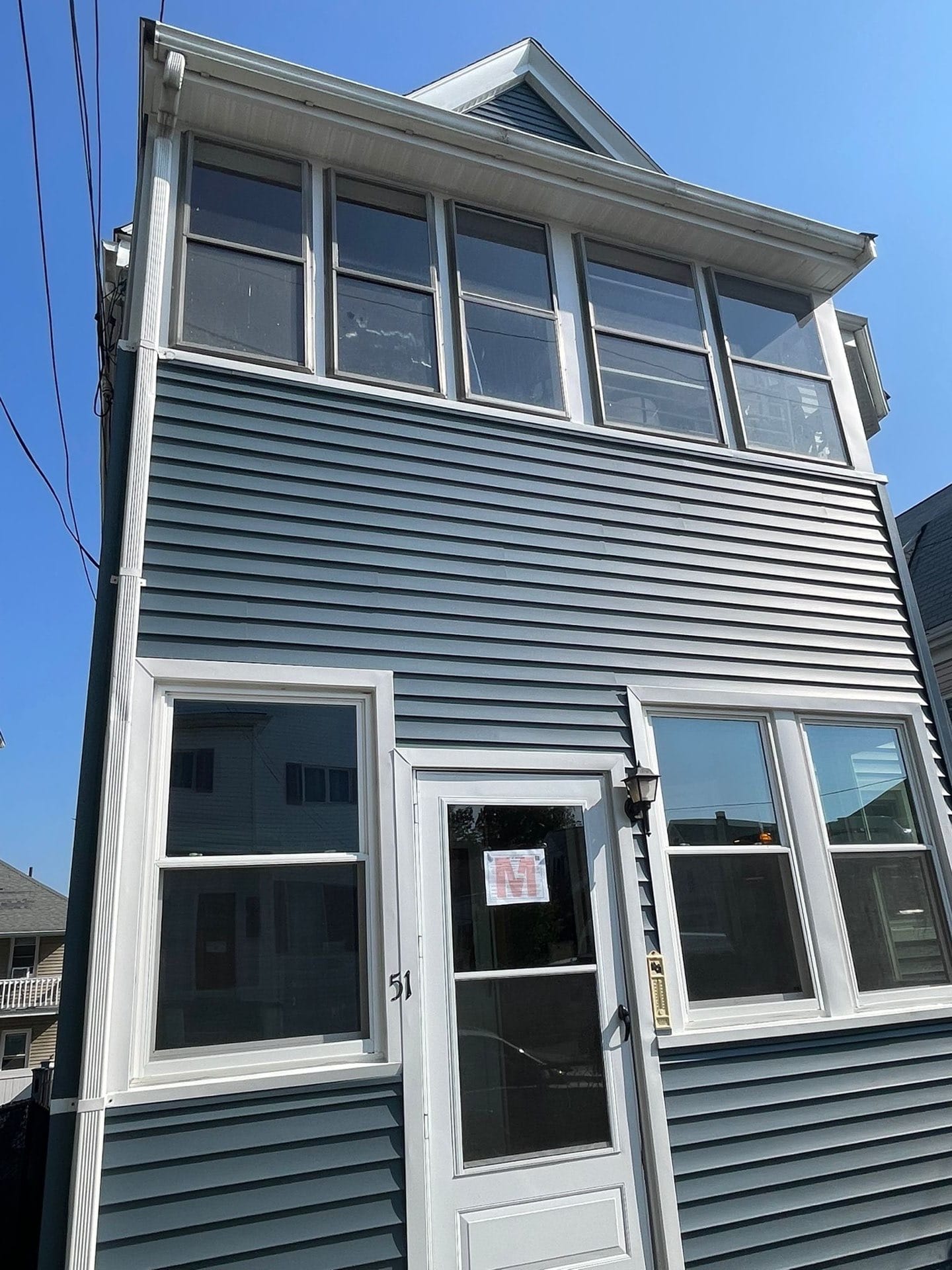 2025-06-05 13 32 32Z-mbjf1tckkxdk3xgl Front facade of a narrow, two-story building with dark blue horizontal siding, white trim, a glass storm door, and a top-floor sun porch.