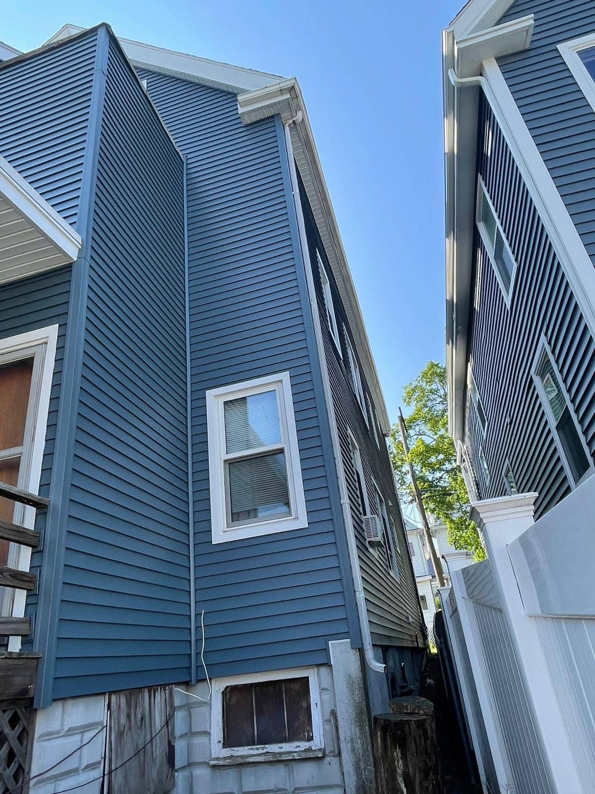 2025-06-05 13 32 32Z-mbjf1tcthwttcoxu View between two closely built houses with dark blue siding and white trim, focusing on the exterior details against a clear blue sky.