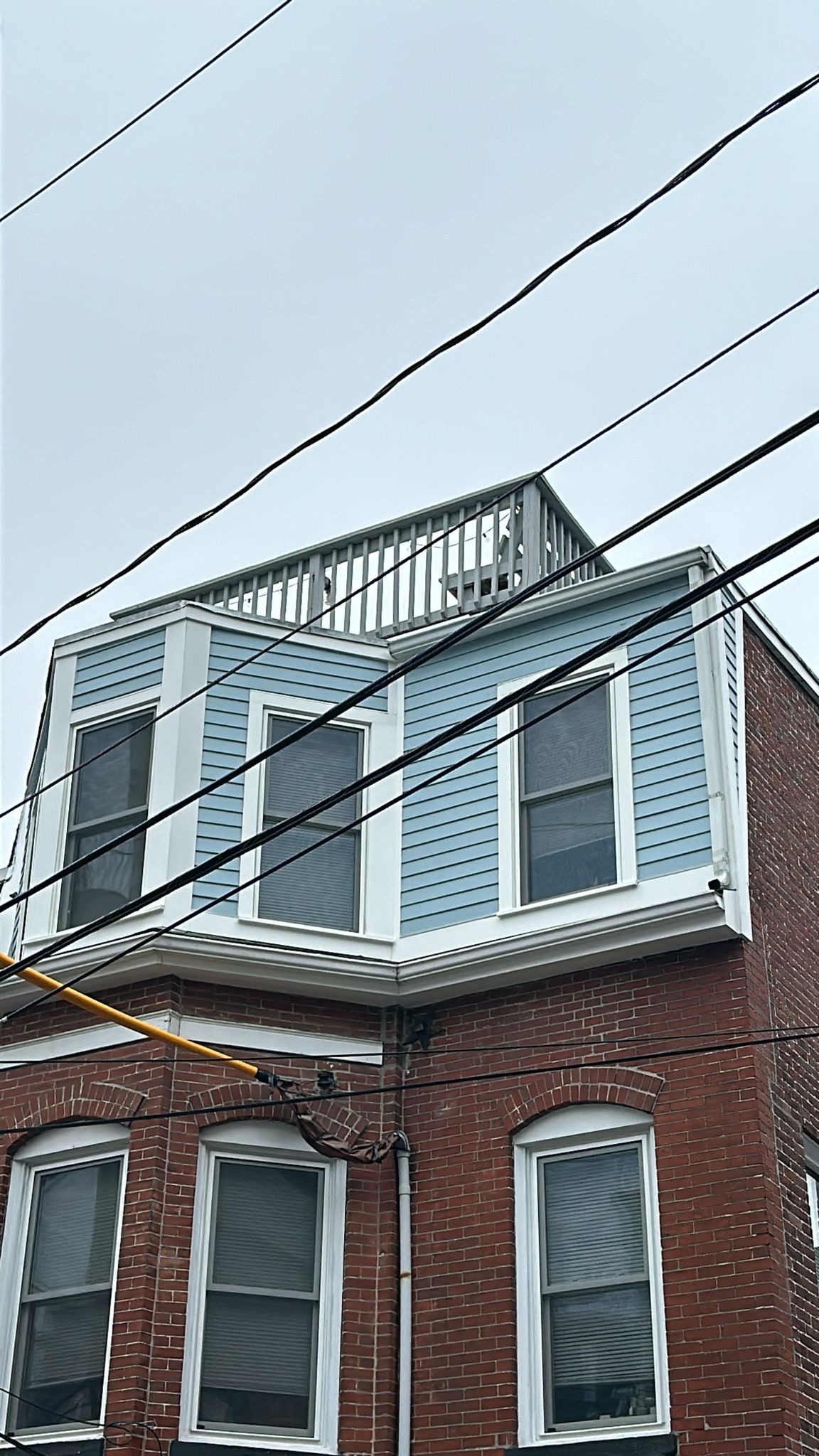 2025-06-10 15 03 04Z-mbqnhi8rmt3uqpq6 Street view of a three-story brick apartment building with bay windows and a mansard roof on an overcast day.