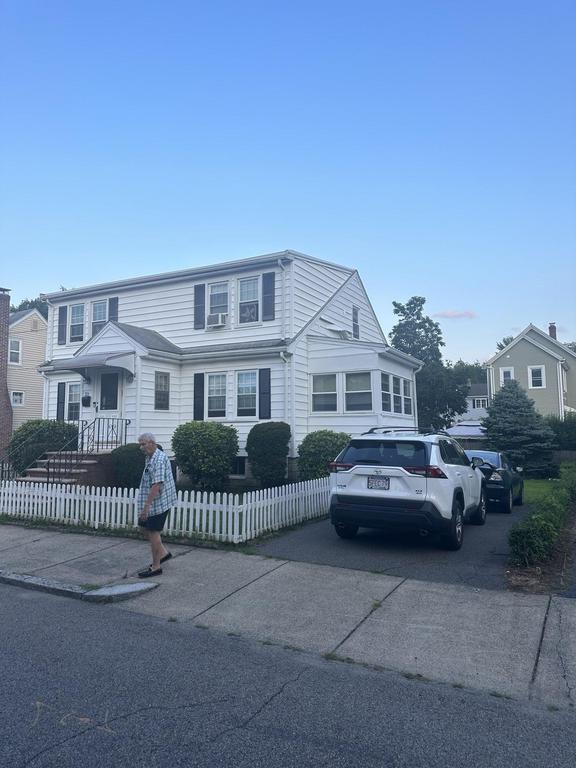 7 24 2023 9 10 20 PM-lkhlo86lcwmri562 White house with black shutters, a white picket fence, and a person walking on the sidewalk.