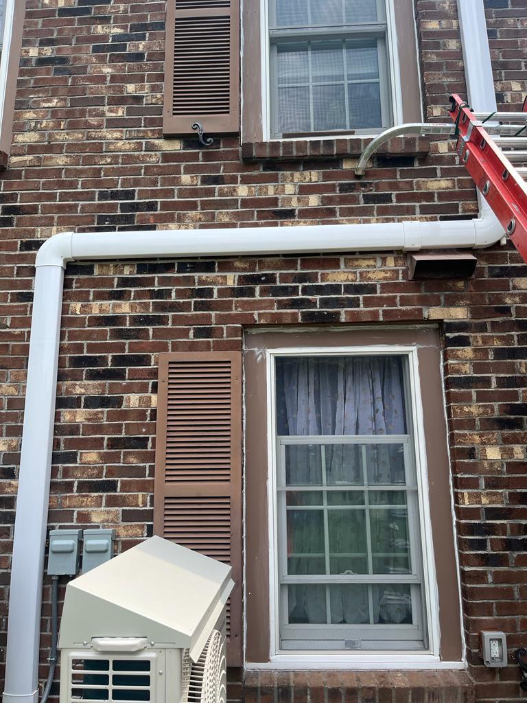 IMG-20230804-WA0005-lkwt5rxrg5wv3oyg a brick wall showing two windows with brown trim and shutters, a white downspout and gutter, and an HVAC unit cover below.