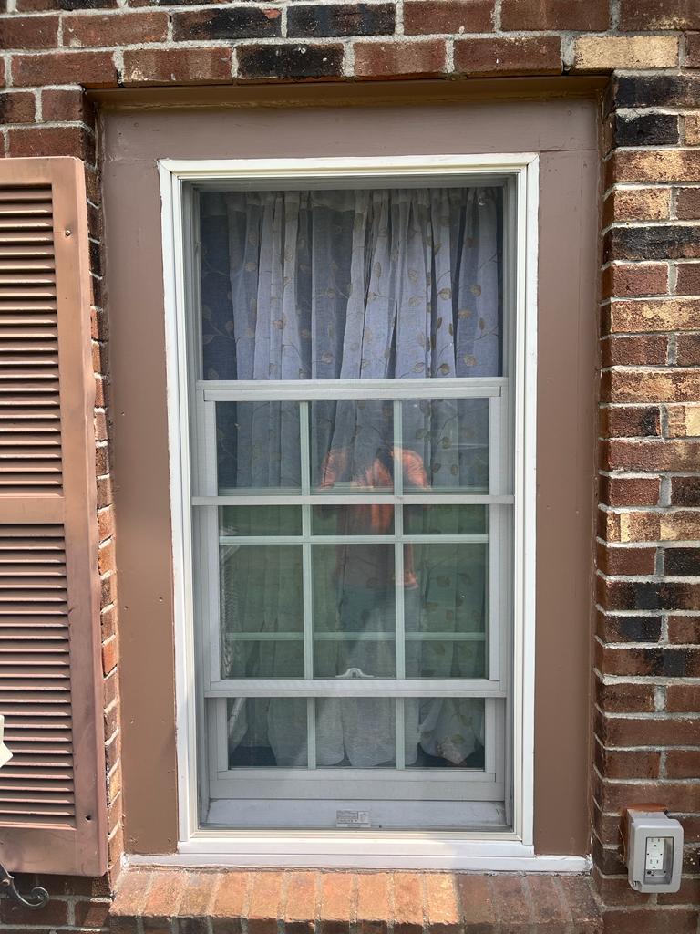 IMG-20230804-WA0007-lkwt5rx4en4qfw7f a double-hung window with a grid pattern, framed by dark brown trim against a brick wall, with curtains visible inside.