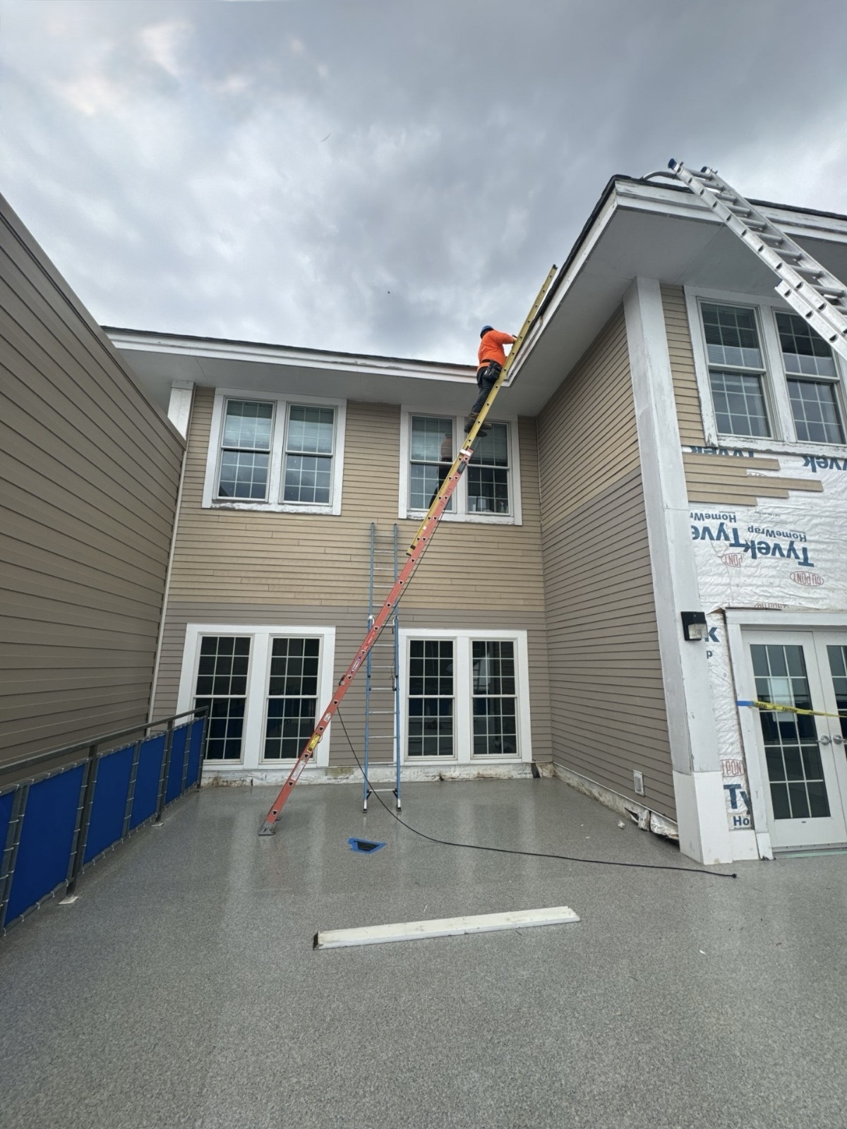 IMG_3695 Residential exterior with tan siding, multiple windows, a grey speckled deck, and a worker making repairs.