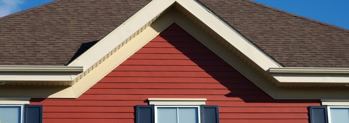 a house exterior featuring a brown shingle roof, red horizontal siding, and a white window with navy blue shutters against a clear blue sky.
