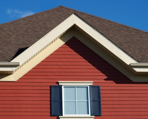 a house exterior featuring a brown shingle roof, red horizontal siding, and a white window with navy blue shutters against a clear blue sky.