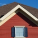 a house exterior featuring a brown shingle roof, red horizontal siding, and a white window with navy blue shutters against a clear blue sky.